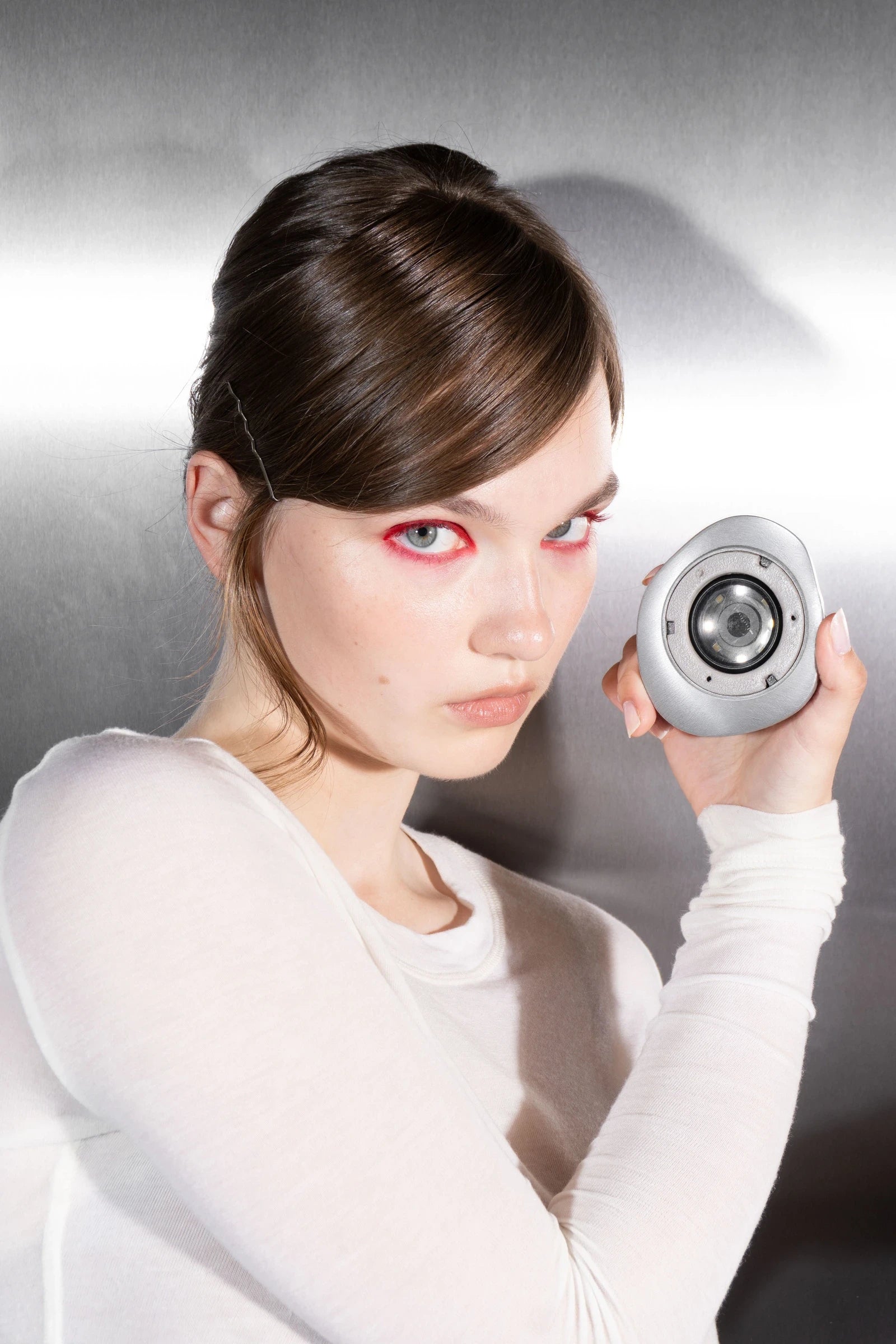A model with red eyeliner and a white top holds up the open Rhia Skin tracker and its lens at a chrome wall