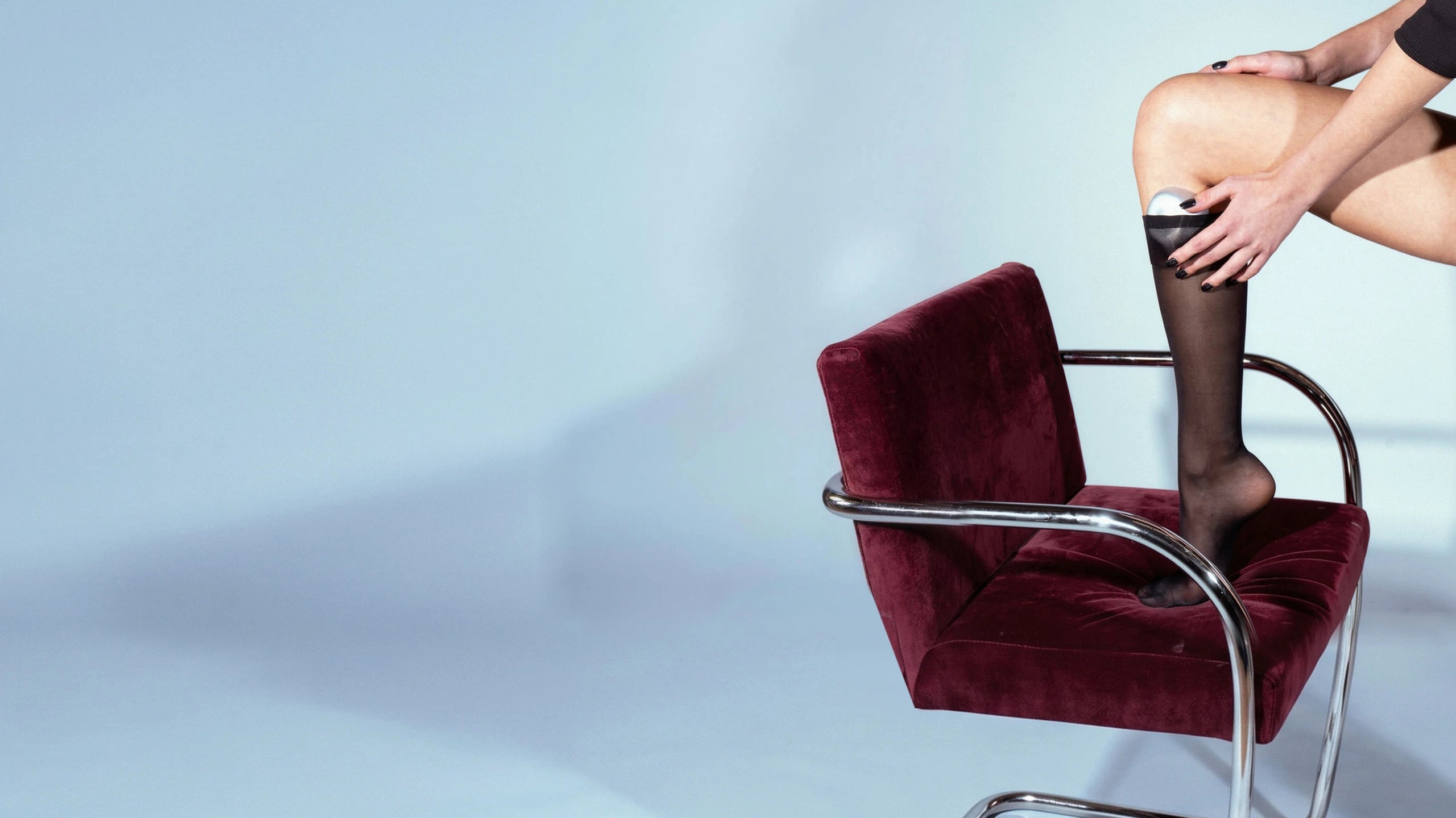 A model in sheer black socks holds the silver Rhia Skin tracker to the back of her knee, one foot on a red velvet chair with chrome arms