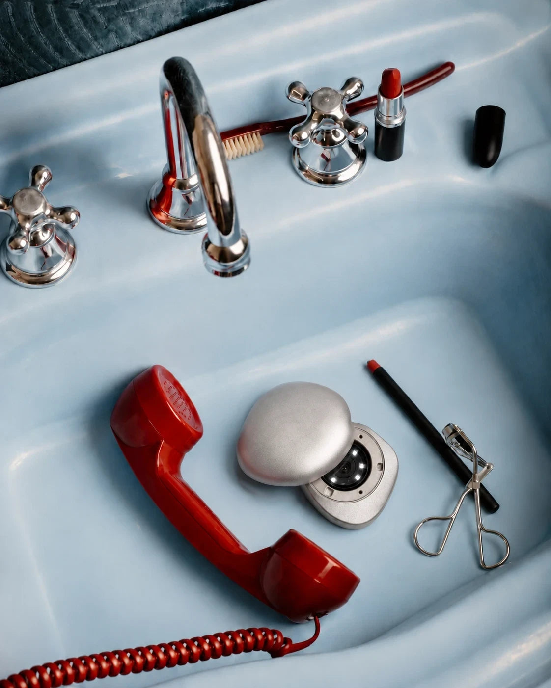 The open Rhia Skin tracker in a pale blue sink beside a red telephone receiver, red lipstick, a lip pencil and an eyelash curler